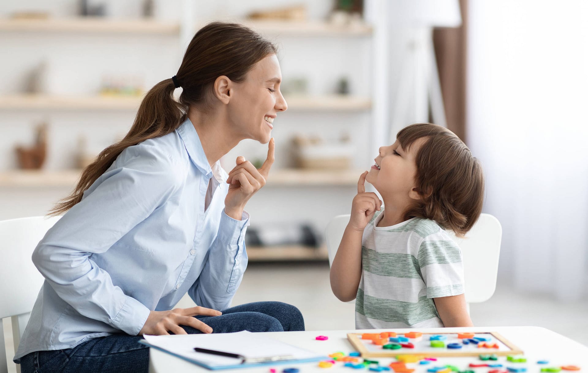 Speech training for kids. Professional woman training with little boy at cabinet, teaching right articulation exercises Speech training for kids. Professional woman specialist training with little boy at cabinet, teaching him right articulation exercises, side view