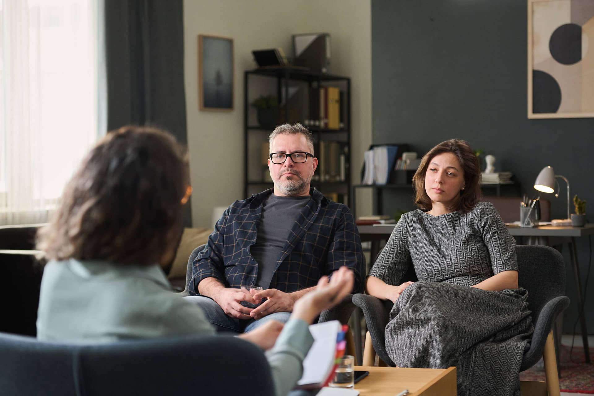 Discussing Relationship Issues in Couple Therapy Session Couple sitting in office discussing relationship issues with therapist holding clipboard. Room has bookshelf, lamp, abstract painting, and large window providing natural light